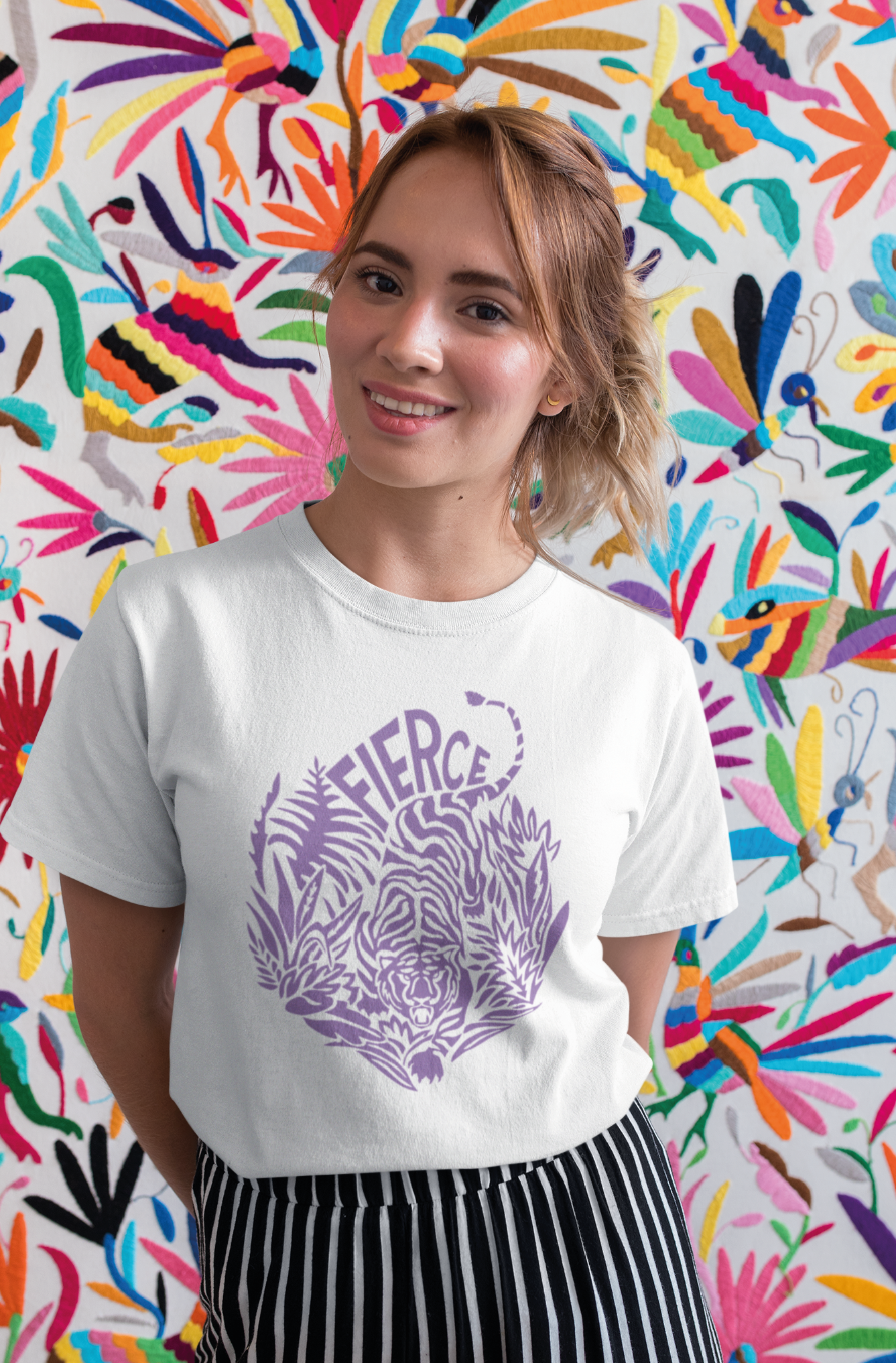 Woman standing in front of a patterned and colorful backdrop smiles while wearing a white tshirt with a Braver Tiger print in lavendar