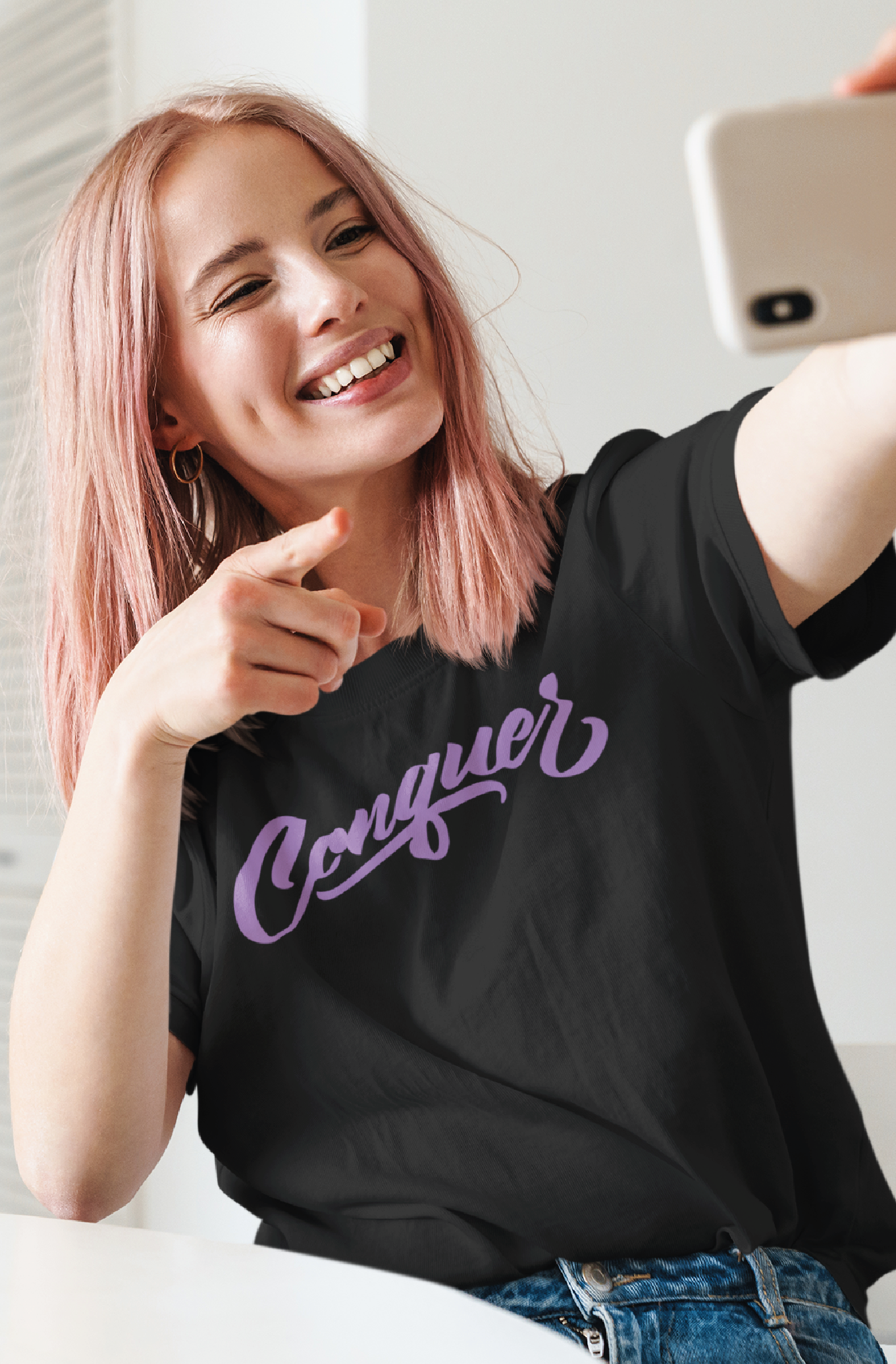 Women with pink hair taking a selfie wearing a black t-shirt with the word conquer printed in lilac on the chest
